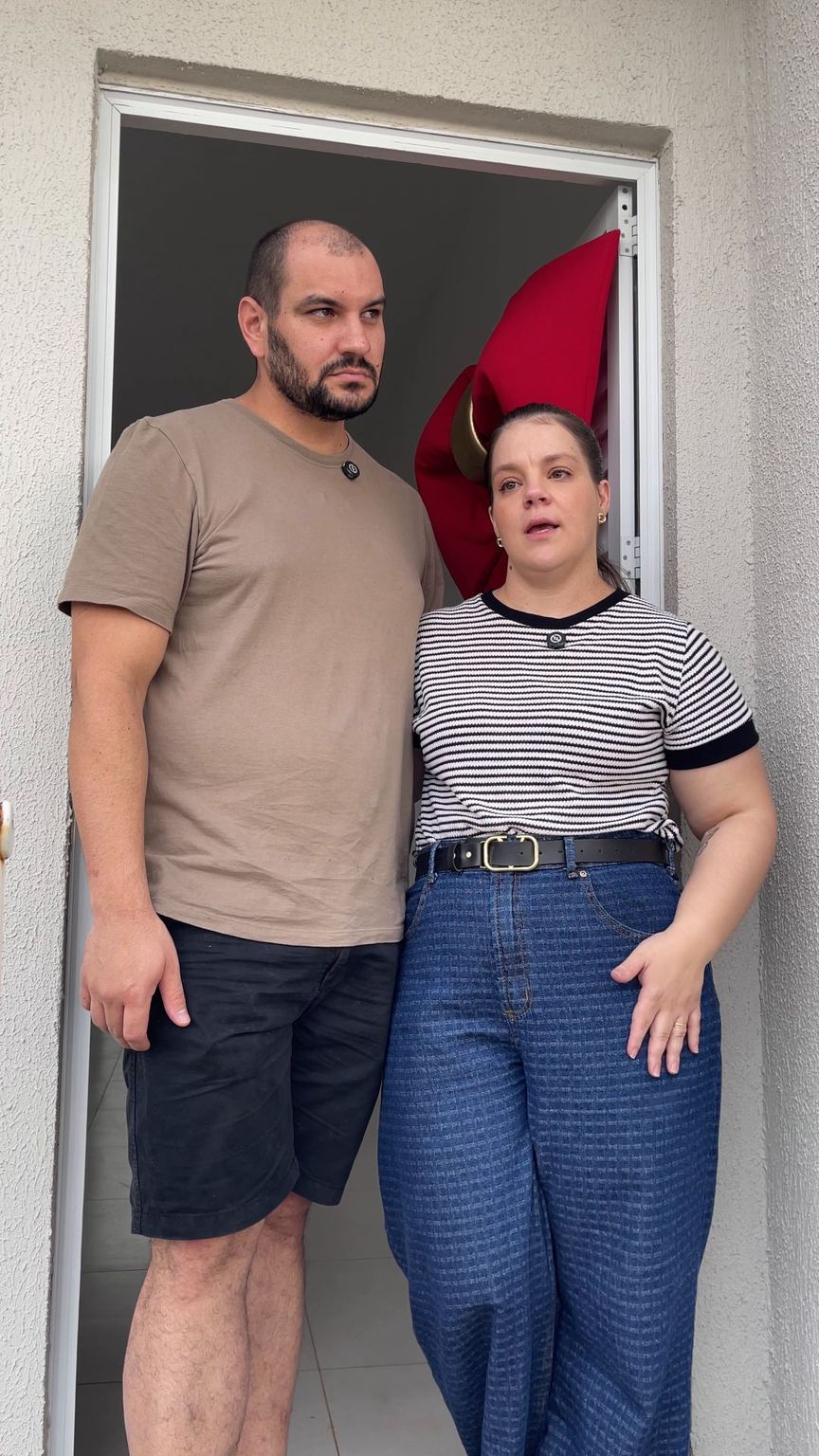 Casal sorri na porta da casa decorada com laço vermelho.