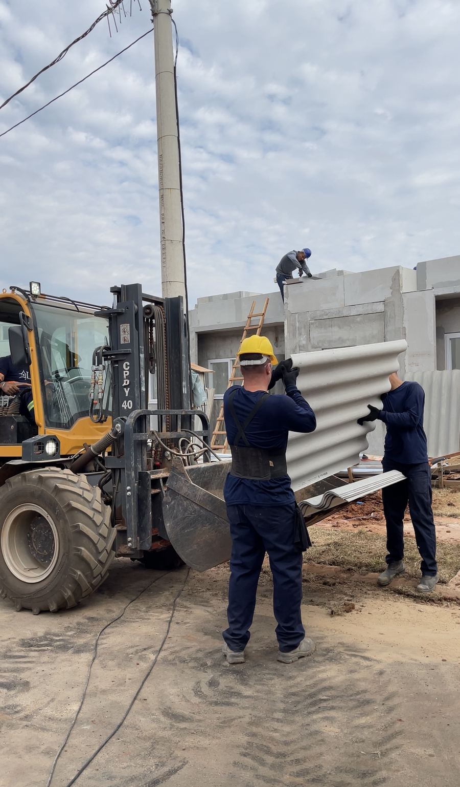 Equipe de capacete descarrega material na obra com a ajuda de uma máquina.