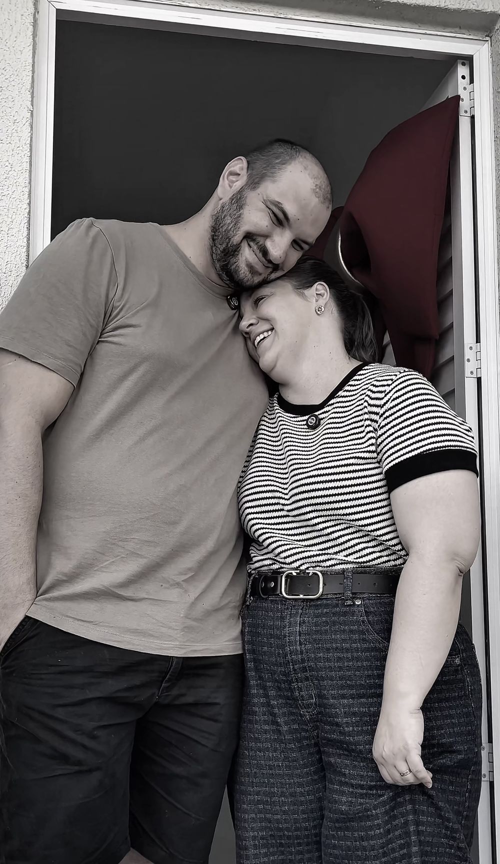 Casal abraçado na porta da casa nova, com o laço vermelho atrás.